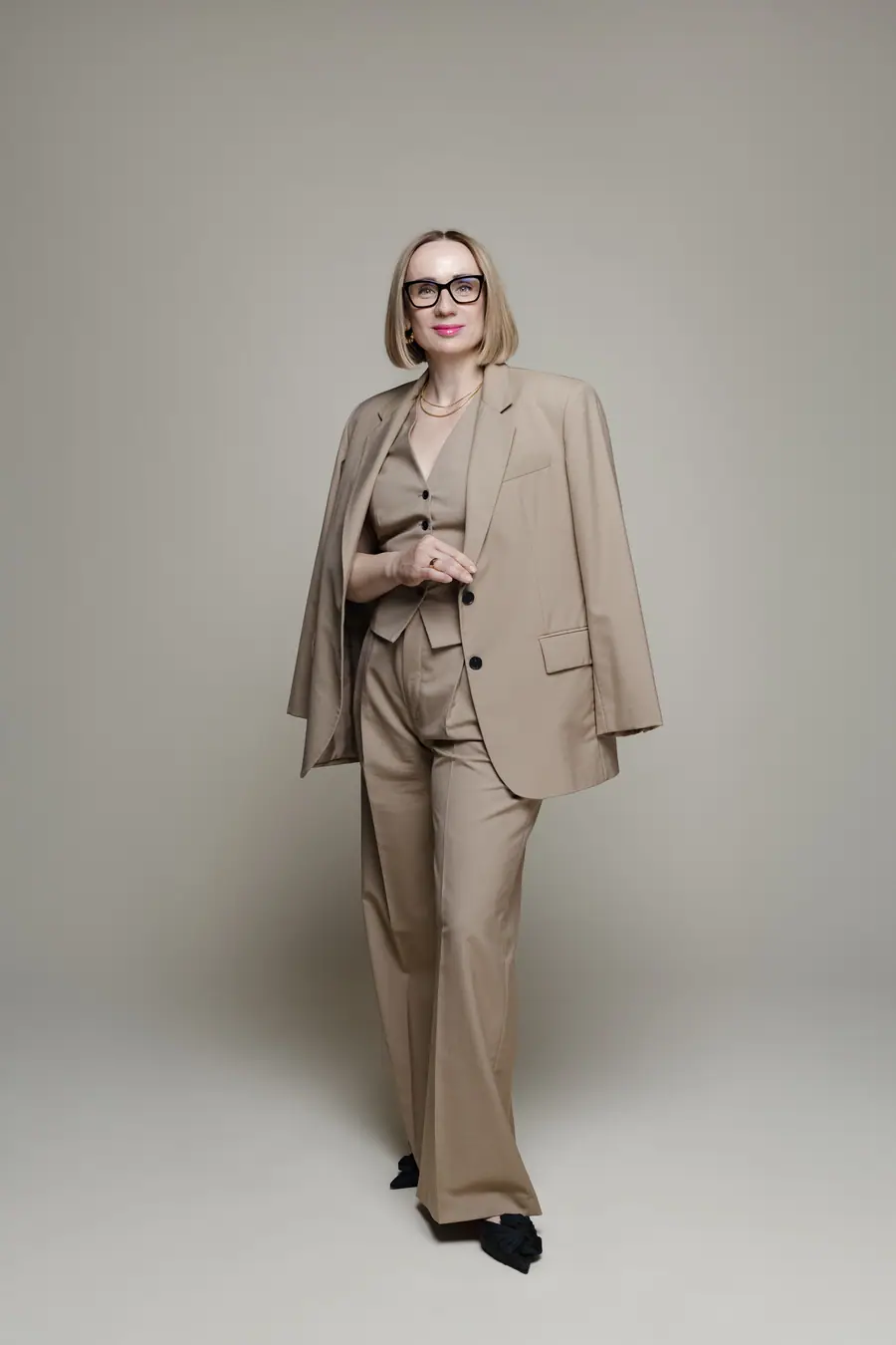 Woman in a beige suit standing full length against a plain wall