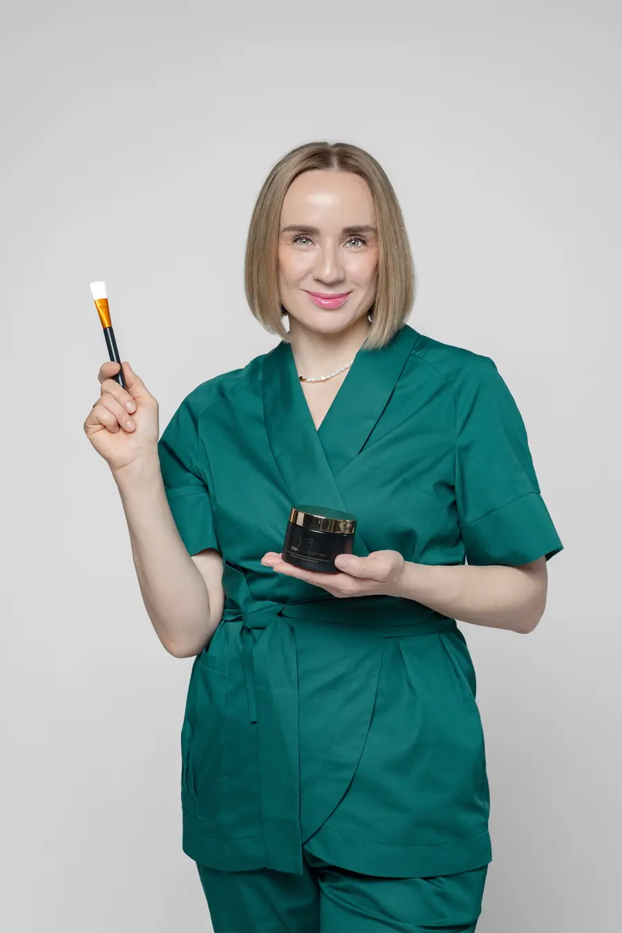 Woman in green scrubs holding a jar of cream and a mask brush