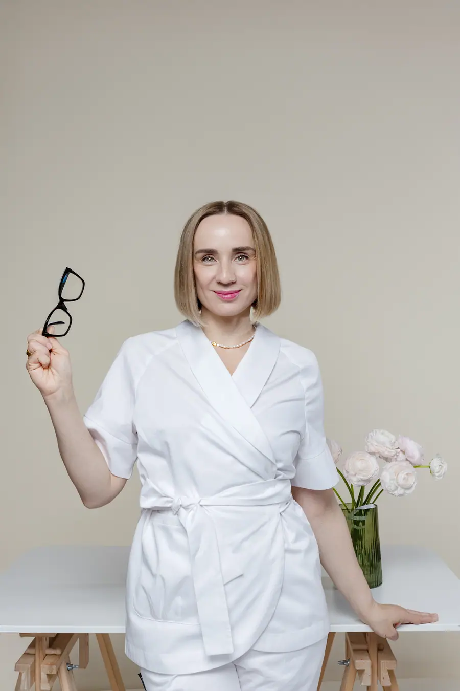 Woman in a white uniform holding her glasses, beside a vase of flowers